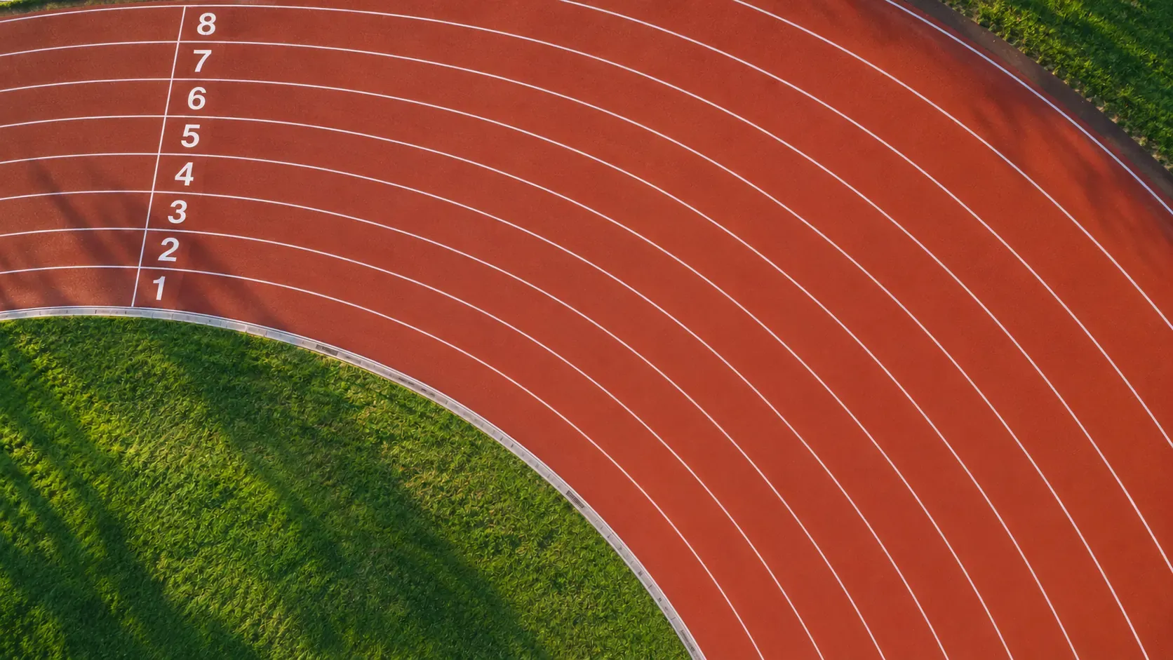 An aerial view of the bend of a red running track, its white lane lines and lane numbers, in early morning light
